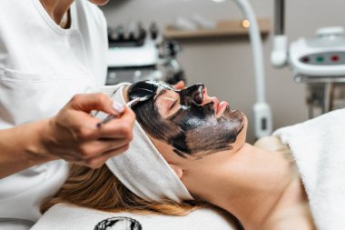 Kozmetikçi, genç bir kadının yüzüne, profesyonel bir spa ortamında cildini geliştirmek için fırça kullanarak canlandırıcı bir siyah maske takıyor.