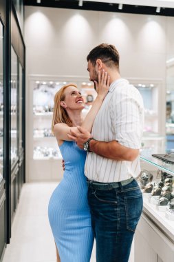 Parlak bir mağazadan mücevher alırken birbirlerine sarılıp gülümseyen mutlu çift, kolye ve saat seçerken birlikte özel bir anın tadını çıkarıyorlar.