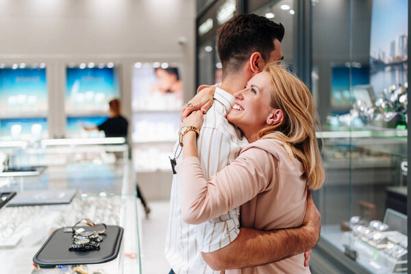 Happy couple hugging and smiling at each other while shopping for jewelry in a brightly lit store, enjoying a special moment together as they browse the selection of necklaces and watches
