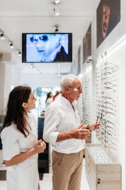 Gülümseyen kadın gözlükçü, modern bir optik mağazada yeni gözlük seçen yaşlı bir adama yardım ediyor, profesyonel tavsiyeler veriyor ve özel hizmet veriyor.