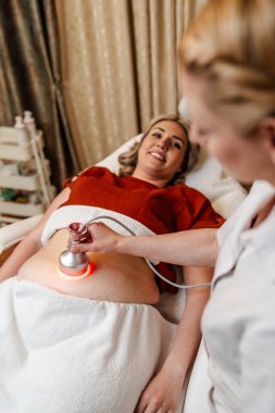 Modern güzellik merkezinde müşterinin karnına kırmızı ışık tedavisi uygulayan profesyonel güzellik uzmanı cilt gençleştirme ve kolajen üretimini teşvik ediyor.