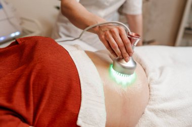 Modern güzellik merkezinde müşterinin karnına yeşil ışık terapisi uygulayan profesyonel güzellik uzmanı cilt gençleştirme ve kolajen üretimini teşvik ediyor.