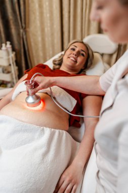Modern güzellik merkezinde müşterinin karnına kırmızı ışık tedavisi uygulayan profesyonel güzellik uzmanı cilt gençleştirme ve kolajen üretimini teşvik ediyor.
