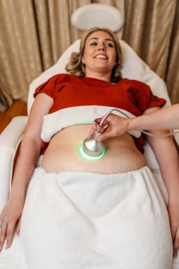 Modern güzellik merkezinde müşterinin karnına yeşil ışık terapisi uygulayan profesyonel güzellik uzmanı cilt gençleştirme ve kolajen üretimini teşvik ediyor.