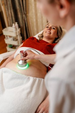 Modern güzellik merkezinde müşterinin karnına yeşil ışık terapisi uygulayan profesyonel güzellik uzmanı cilt gençleştirme ve kolajen üretimini teşvik ediyor.