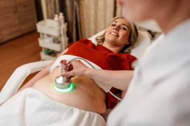 Modern güzellik merkezinde müşterinin karnına yeşil ışık terapisi uygulayan profesyonel güzellik uzmanı cilt gençleştirme ve kolajen üretimini teşvik ediyor.