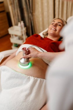 Modern güzellik merkezinde müşterinin karnına yeşil ışık terapisi uygulayan profesyonel güzellik uzmanı cilt gençleştirme ve kolajen üretimini teşvik ediyor.