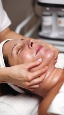 Profesyonel güzellik uzmanı olgun bir kadının boynuna masaj yapıyor. Güzellik merkezinde bir tedavi yatağında yatıyor. Arka planda modern ekipmanlar sayesinde rahatlatıcı ve canlandırıcı bir deneyim yaşıyor.