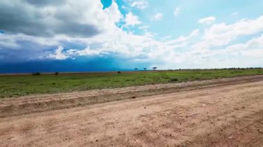 Geniş açık gökyüzü ve çimenli arazide ilerleyen toprak yolu olan Afrika düzlükleri. Uzakta fırtına kopuyor..