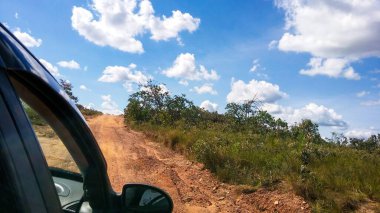 Bir araba penceresinden panoramik bir manzara. Bereketli bir bitki örtüsüyle çevrili engebeli bir toprak yolu yakalıyor. Tüylü beyaz bulutlarla kaplı, canlı mavi bir gökyüzü..