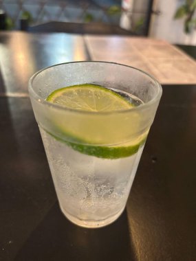 Canlı yeşil limon dilimleriyle kaplı ferahlatıcı karbonatlı bir içecekle dolu kristal berrak bir bardak. Aydınlıkta parıldayan içki, bulanık bir arka planı olan karanlık bir masaya kurulmuş, rahatlamış bir atmosfer yayıyor sosyalleşmek için mükemmel ya da değil.