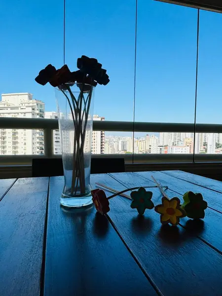 Parlak mavi bir gökyüzüne ve büyük pencerelerden görünen şehir siluetine karşı duran temiz cam bir vazoda renkli kağıt çiçek çubukları. Ahşap masa bu neşeli ve canlı iç mekan sahnesine kırsal bir unsur ekliyor.