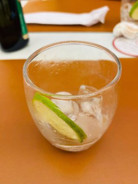 İçinde buz ve bir dilim limon olan temiz bir bardak su ya da karbonatlı bir içecek. Görüntünün arka planı, açık turuncu masa örtüsü ve görünür çatal bıçakları olan bir restoran masasını gösteriyor..