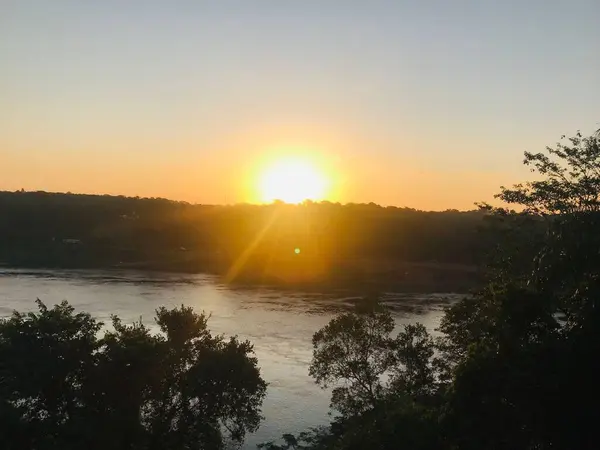 Güzel bir günbatımı, nehrin sakin suları üzerinde turuncu ve sarı renkleri yansıtan ufku aydınlatır. Nehir kıyısındaki ağaçların siluetleri huzur ve sükunet hissi katar, rahatlama ve tefekkür için mükemmel bir sahne yaratır. 