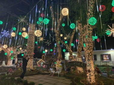 Ağaçlara renkli ışıklar asılan süsler ve tatilin keyfini yakalayan büyülü bir atmosferle dolu büyüleyici bir Noel sahnesi. Çocuklar arka planda oynar ve gülümserken yıldız şeklinde ışıklar ve pırıltılı mücevherler...