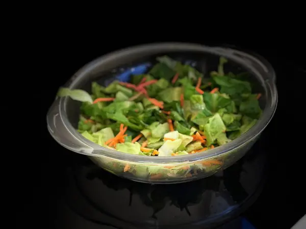 Koyu bir zemin üzerinde taze yeşil yapraklar ve havuç parçalarından oluşan taze bir salatayla dolu temiz bir kâse içeriğinin canlılığını ve tazeliğini vurguluyor..