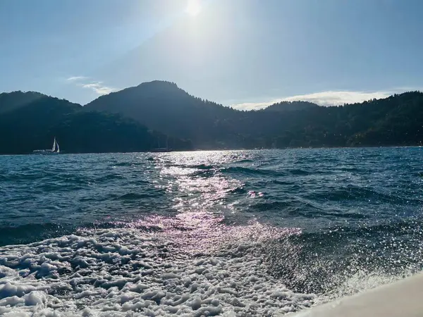 Güneş ışığını yansıtan okyanus dalgalarını yakalayan sakin bir sahnenin görüntüsü. Arka planda, yeşil dağlar berrak mavi gökyüzüne karşı yükselirken, küçük bir yelkenli sakin bir şekilde köpüklü sularda yüzüyor..
