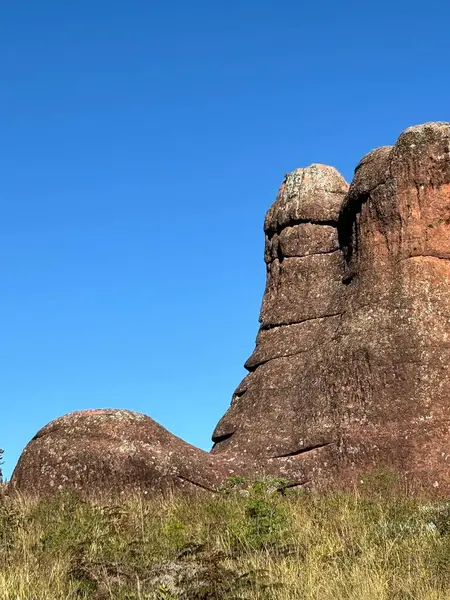 Görkemli bir kaya oluşumu açık mavi gökyüzüne karşı duruyor. Mor ve kahverengi dokular yemyeşil doğanın ortasında büyüleyici bir manzara yaratıyor. Doğal ışık kayayı aydınlatır, benzersiz şekillerini ve etrafındaki vahşi çevreyi vurgular. 