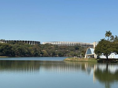 Güneşli bir günde ünlü stadyum ve modern mimariyi aydınlatan, etrafını çevreleyen ağaçların yansımasıyla panoramik bir göl manzarası. Sahne, doğa ve şehirleşme arasındaki uyumu yakalar..
