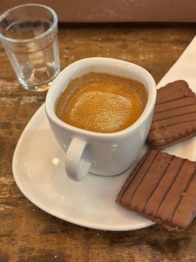 Köy ahşap bir masada servis edilen bir fincan taze espresso. Yanındaki iki güzel dekore edilmiş çikolatalı kurabiye kahvenin canlı tonuyla tezat oluşturuyor. Bir bardak berrak su bileşimi tamamlar..