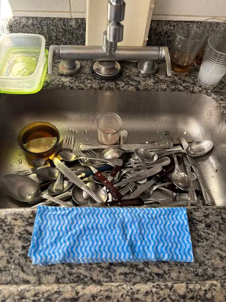 Kirli tabaklarla dolu bir mutfak lavabosu, gümüş takımlar, bardaklar ve ön planda mavi bir masa örtüsü olan bir kase. Bu sahne bir aile yemeğinden sonraki bir anı hatırlatıyor. Su ve tabaklar w 'dan gelen doğal ışığı yansıtıyor.