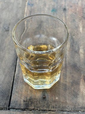 Kırsal bir ahşap masada üzerinde altın bir içecek bulunan berrak bir bardak. Işık sıvının yüzeyinden nazikçe yansıyarak sıcak ve davetkar bir atmosfer yaratır, rahatlatıcı anlar için mükemmeldir..