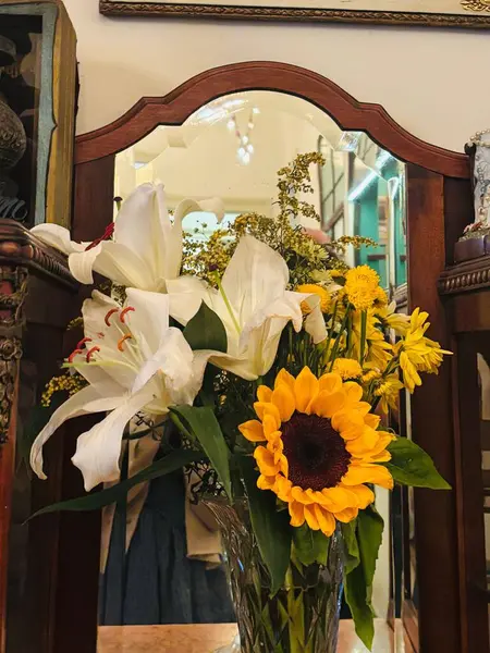 Beyaz zambaklar, canlı ayçiçekleri ve kırsal bir masada sarı papatyalar içeren çeşitli çiçeklerle dolu güzel bir vazo. Düzenleme arka planda dekoratif bir aynaya yansıyarak sıcak ve neşeli bir atmosfer yaratıyor..