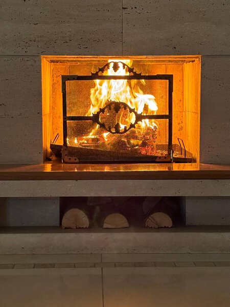 An intimate and cozy view of a lit fireplace, with dancing flames illuminating a crackling fire that brings warmth and comfort to the space. The metal structure contrasts with natural stones, while stacked firewood adds a rustic touch, perfect for ev