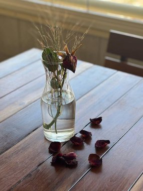 Koyu ahşap bir masanın üzerine yerleştirilmiş, temiz cam vazoda kurumuş çiçeklerin hassas bir düzenlemesi. Etrafa saçılmış kurumuş gül yaprakları ve pencereden gelen doğal ışıkla aydınlatılmış bir arka plan..