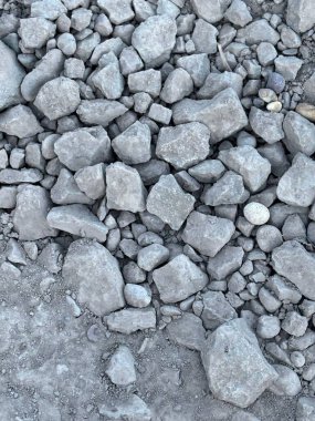 Gri taşlar ve çakıllarla kaplı bir yüzeye yakın plan, çeşitli boyutlarda ve şekillerde. Kabataslak ve pürüzsüz dokular arasındaki zıtlık sahnenin doğallığını ve paslanmışlığını gösterir..