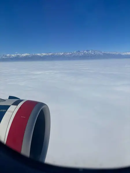 Bir uçak penceresinden etkileyici bir hava manzarası, altında pamuk gibi geniş beyaz bulutlar sergiliyor, arkaplanda görkemli bir kar örtülü dağ aralığı açık mavi bir gökyüzünün altında..