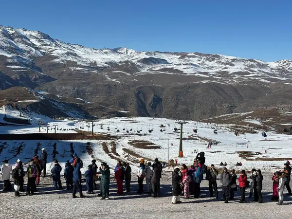 Güneşli bir günde, karlı bir dağda bir grup insan manzaranın tadını çıkarıyor. Arka plan, karla kaplı engin dağları ve yeşil bir düzlüğü gösteriyor. Açık mavi gökyüzü sükunet ekler.