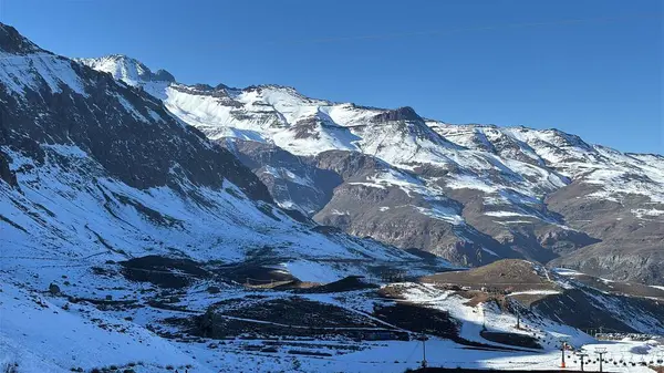 Açık mavi gökyüzünün altında karla kaplı etkileyici bir dağ manzarası. Dik, kayalık yamaçlar, güneşte parlayan kar taneleriyle görülebilir. Huzur ve ihtişamı yansıtan bir sahne, doğa ve macera aşıkları için mükemmel..