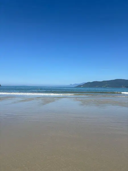 Işığın denizin yumuşak dalgalarıyla buluştuğu sakin bir sahilin çarpıcı manzarası. Açık mavi gökyüzü güneşli bir günü yansıtıyor, sakin ve rahatlatıcı bir atmosfer yaratıyor. Arka plandaki dağlar manzarayı tamamlıyor, barış hissi uyandırıyor.