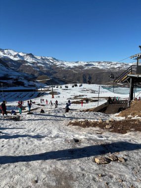 Görkemli dağlarla çevrili insanların karın tadını çıkardığı canlı bir kayak merkezi görüntüsü. Mavi gökyüzü, kayakçılar yamaçlardan aşağı inerken ve diğerleri manzarayı izlerken parlıyor. Atmosfer enerjik ve kış heyecanıyla dolu..