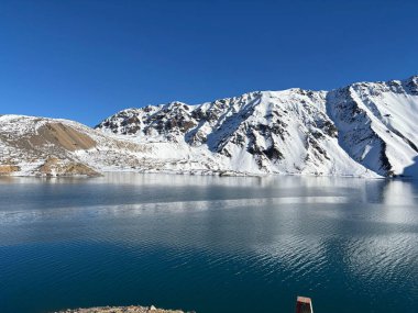 Karla kaplı güzel bir dağda donmuş bir gölün çarpıcı manzarası. Açık mavi gökyüzü sakin sulara yansıyarak sakin ve sakin bir atmosfer yaratır. Yükselen dağlar ihtişam ve doğal güzellik ekler..