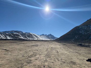 Uçsuz bucaksız bir dağ manzarası, parlak mavi bir gökyüzünün altında karla kaplı tepeleri olan kurak ama çarpıcı bir manzarayı gözler önüne seriyor. Güneş yoğun bir şekilde parlıyor, vahşi güzelliği arttıran büyülü ışınlar saçıyor..