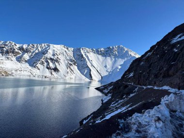 Güneşli bir günde, kristal berrak bir gölün suyuna yansıyan karla kaplı dağların etkileyici bir manzarası. Dik, kayalık yamaçlar beyaz battaniyenin yumuşaklığıyla tezat oluşturur, doğa ve sükunet arasında mükemmel bir uyum yaratır..