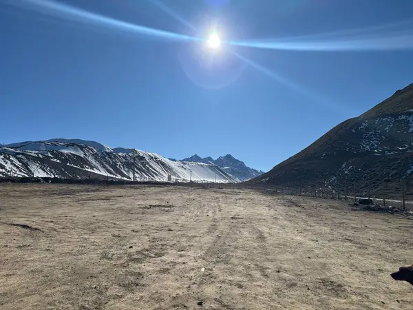 Uçsuz bucaksız bir dağ manzarası, parlak mavi bir gökyüzünün altında karla kaplı tepeleri olan kurak ama çarpıcı bir manzarayı gözler önüne seriyor. Güneş yoğun bir şekilde parlıyor, vahşi güzelliği arttıran büyülü ışınlar saçıyor..