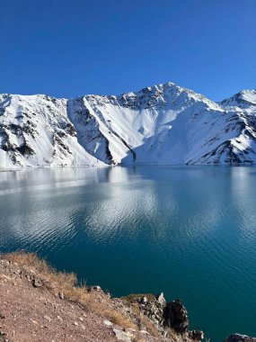 Pırıl pırıl parlayan mavi gökyüzünün altındaki kar kaplı dağları yansıtan berrak bir gölün çarpıcı görüntüsü. Sahne huzurlu bir ortamda dinginlik ve doğal güzelliği çağrıştırıyor..