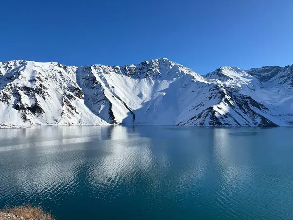 Karla kaplı görkemli dağlarla çevrili donmuş mavi bir gölün etkileyici manzarası. Açık mavi gökyüzü göle yansıyarak huzur ve sükunet ortamı yaratır..