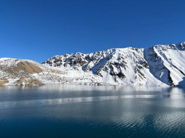berrak bir göl, berrak mavi gökyüzünün altındaki görkemli karla kaplı dağları yansıtıyor. Sahne huzuru ve doğal güzelliği yansıtıyor, dağlık çevrenin saflığını yakalıyor..