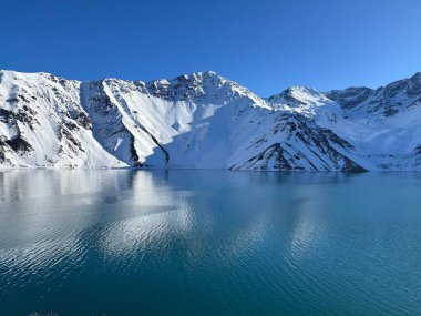 Güneşli bir günde kristal berrak göl, görkemli karlarla kaplı dağlarla çevrili. Sakin sular mavi gökyüzünü ve etkileyici kaya oluşumlarını yansıtıyor. Doğada huzur ve sükunet ortamı yaratıyor..