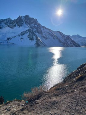 Görkemli karla kaplı dağlarla çevrili sakin bir gölün çarpıcı manzarası. Güneş ışığı suyun üzerinde parıldıyor, parıldayan yansımalar yaratıyor. Manzara, sakin ve izole bir ortamda huzur ve doğal güzelliği çağrıştırıyor..