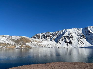 Sakin bir gölün panoramik görüntüsü berrak mavi gökyüzünün altındaki görkemli karla kaplı dağlarla çevrili. Kristal berraklığındaki sudaki yumuşak yansımalar sükuneti ve doğal güzelliği çağrıştırıyor..