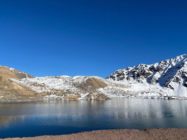 Berrak mavi bir gökyüzünün altında görkemli karla kaplı bir dağ gölü panoramik manzarası. Sudaki yumuşak yansımalar, doğa severler ve gezginler için mükemmel bir sükunet ve doğal güzellik ortamı yaratır..