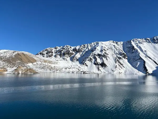 berrak bir göl, berrak mavi gökyüzünün altındaki görkemli karla kaplı dağları yansıtıyor. Sahne huzuru ve doğal güzelliği yansıtıyor, dağlık çevrenin saflığını yakalıyor..