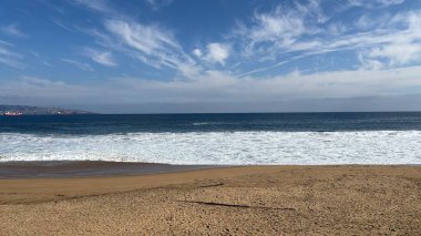 Güneşli bir günde huzurlu bir plajın çarpıcı panoramik manzarası. Dalgalar nazikçe altın kumda kırılıyor, narin bulutlar mavi gökyüzünde yüzerken, huzur verici ve rahatlatıcı bir sahne yaratıyor..