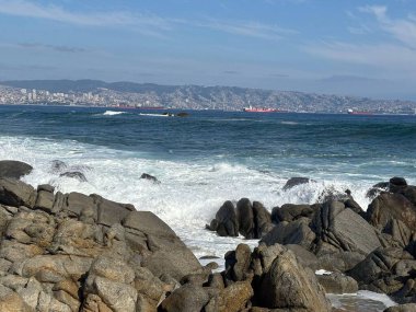 Beyaz dalgaların engebeli kayalara çarptığı sahnenin muhteşem manzarası. Arka planda, kargo gemileri açık bir gökyüzünün altında sakin ve ferahlatıcı bir atmosfer yaratarak, bulutlarla birlikte mavi bir denizde yol alırlar..