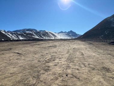 Uçsuz bucaksız kurak arazisi ve parlak mavi gökyüzü ile etkileyici bir çöl manzarası. Arkaplanda karla kaplı dağlar, kuru zemin ve açık ufukla keskin bir tezat oluşturarak görkemli bir şekilde yükseliyor. Yoğun güneş ışığı bir u oluşturur.